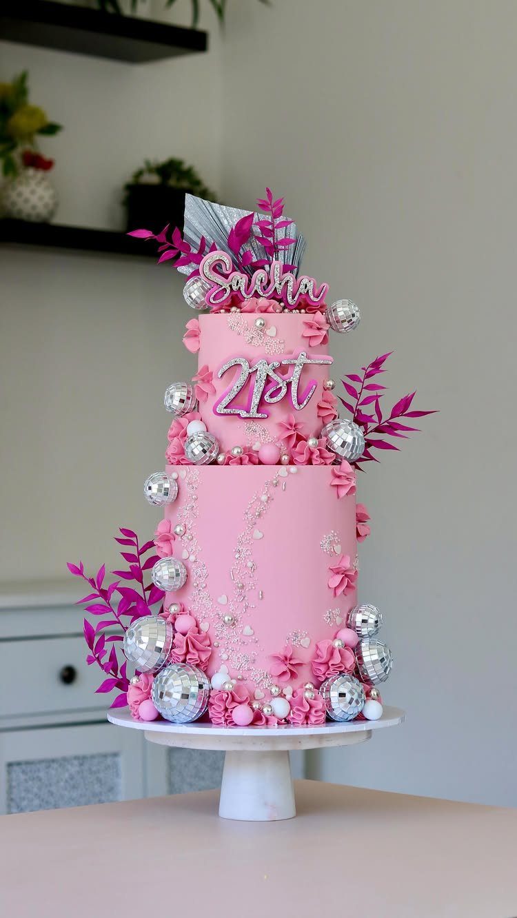 Three-tier pink frosted cake decorated with silver disco balls and 21st birthday toppers.