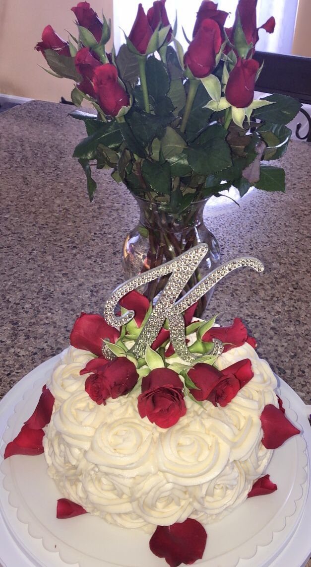 White rosette frosted cake topped with red roses and a silver glittery letter K