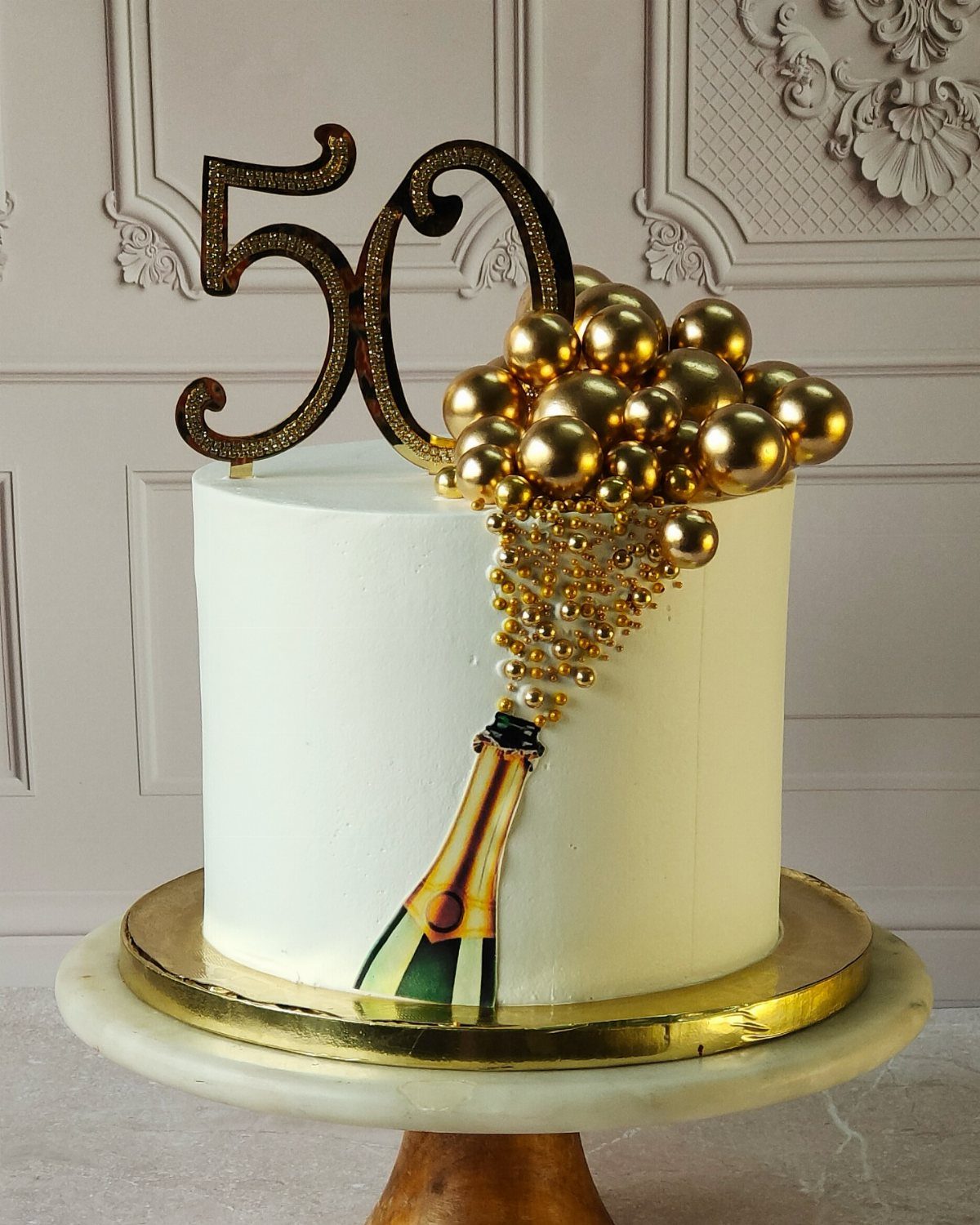 White birthday cake with gold 50 topper, champagne bottle, and gold bubble spheres.