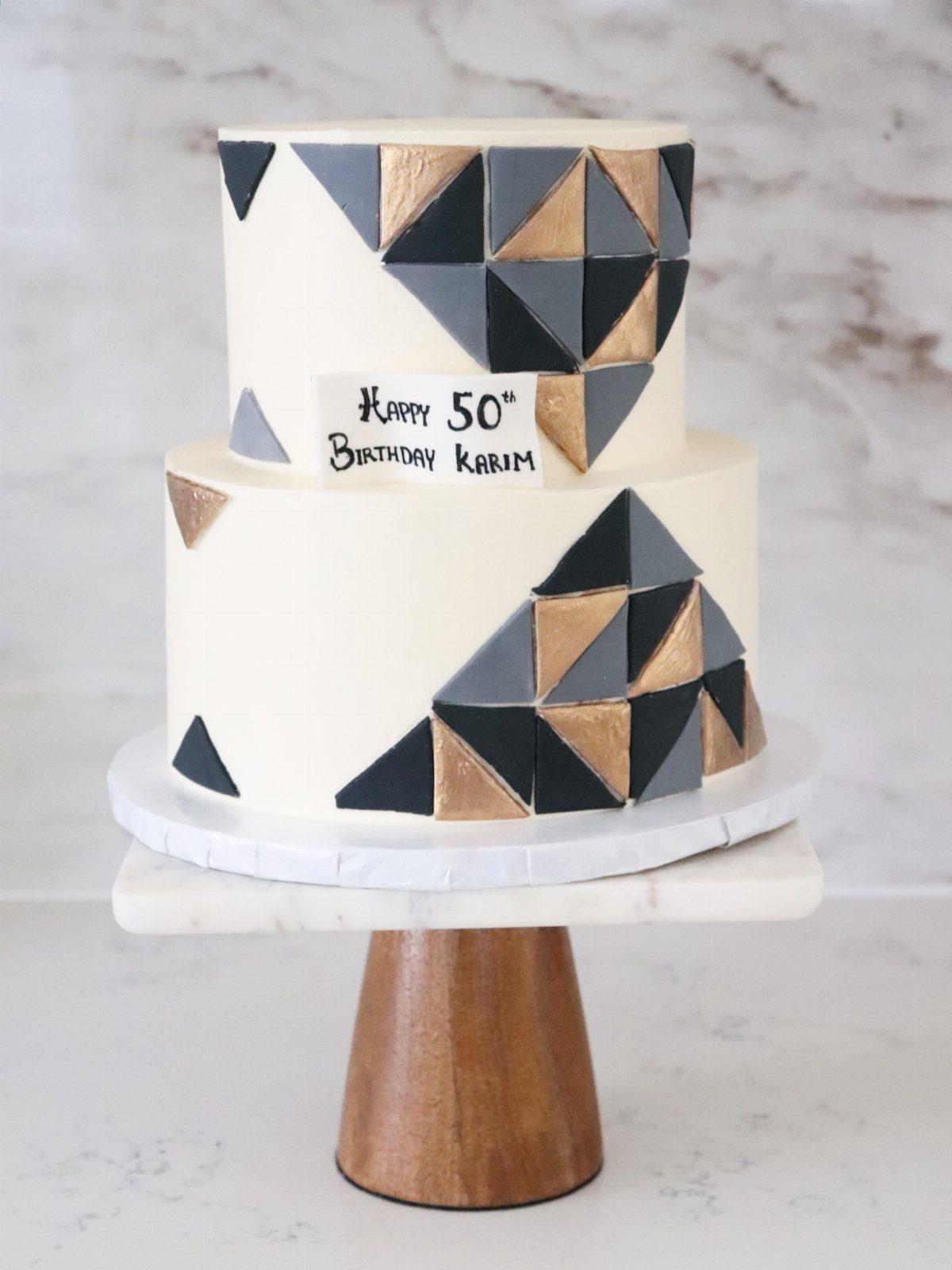 Two-tier white birthday cake decorated with black and gold geometric triangular patterns.