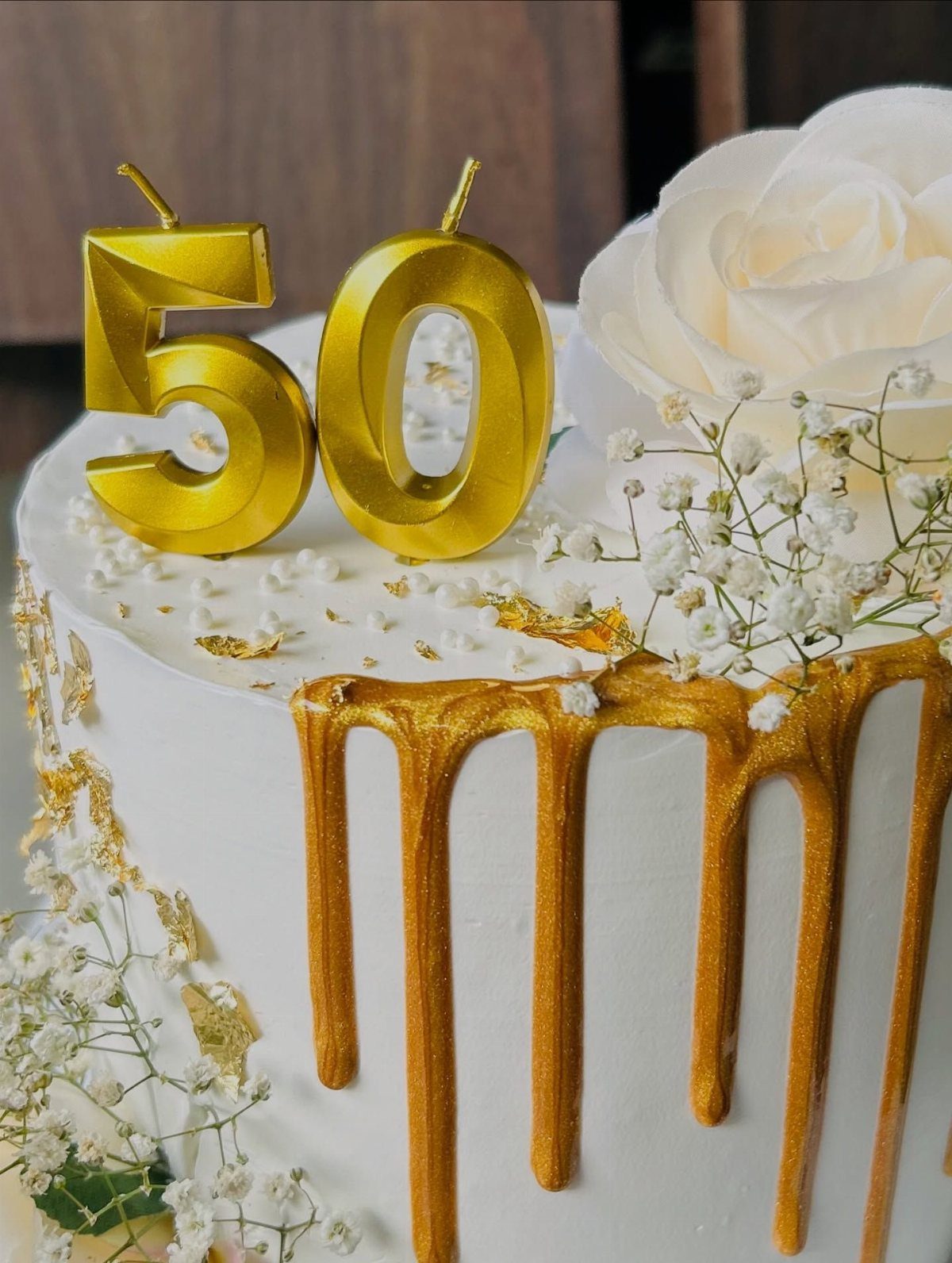 White cake with gold drip, gold 50 candles, and white floral decorations.