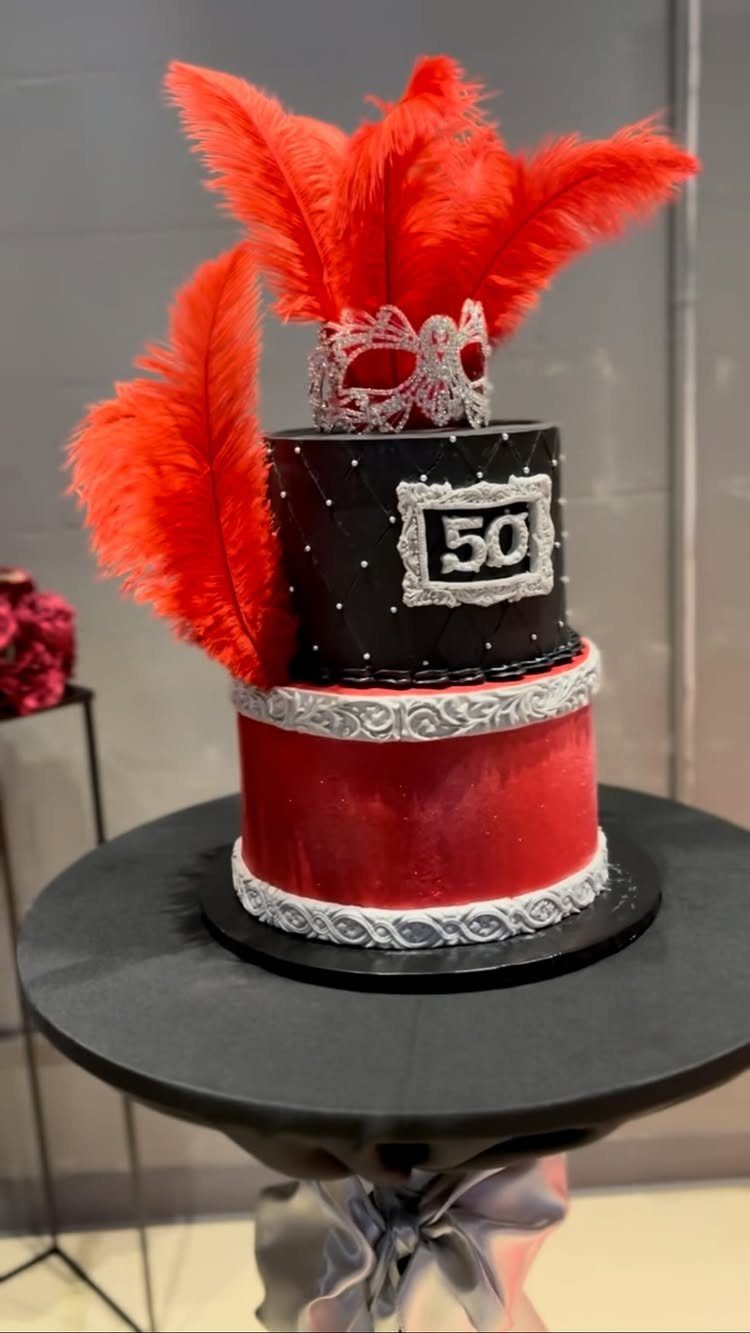 Two-tier red and black cake topped with a silver mask and red feathers.