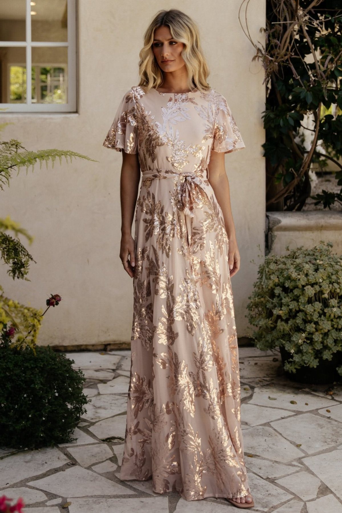 Woman wearing a gold metallic floral print maxi dress on a stone patio