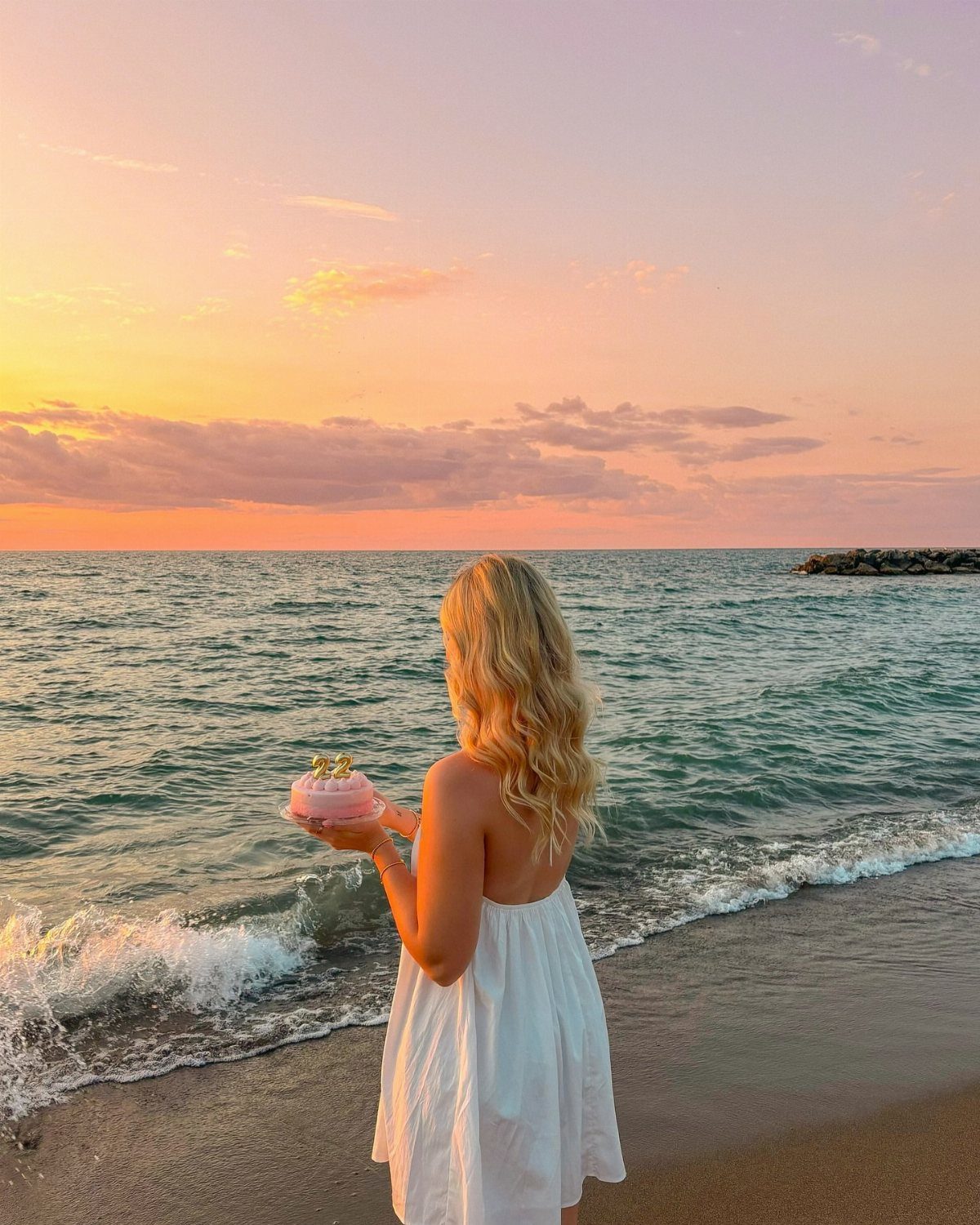 Blonde woman in white dress holding pink cake on a beach at sunset