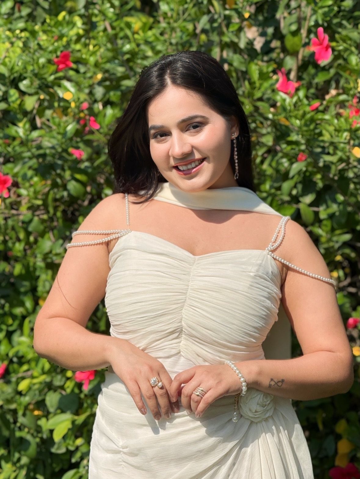 Woman wearing a cream strapless gown with pearl straps posing before green foliage.