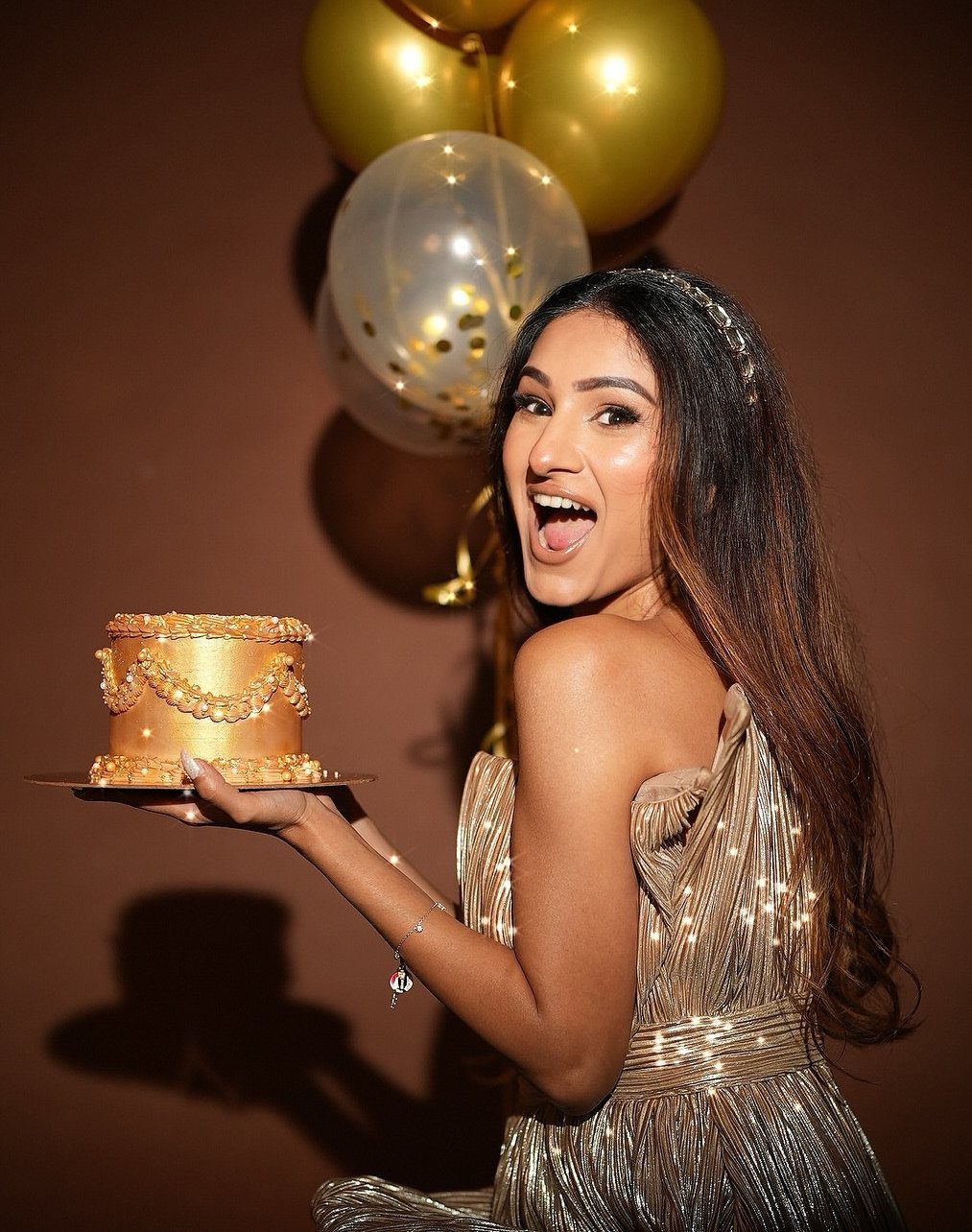 Woman in gold sequin gown holding a gold cake with gold balloons behind.