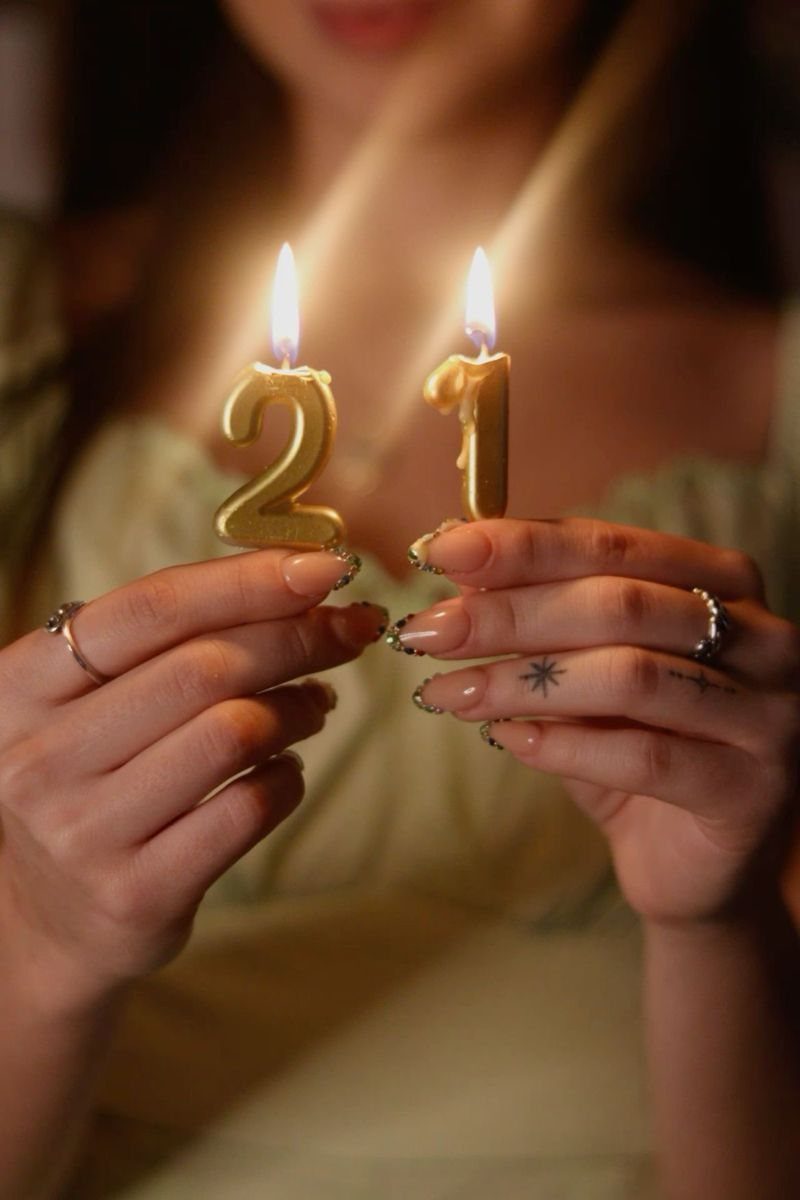 Hands holding lit gold number 21 candles against a blurred cream dress background