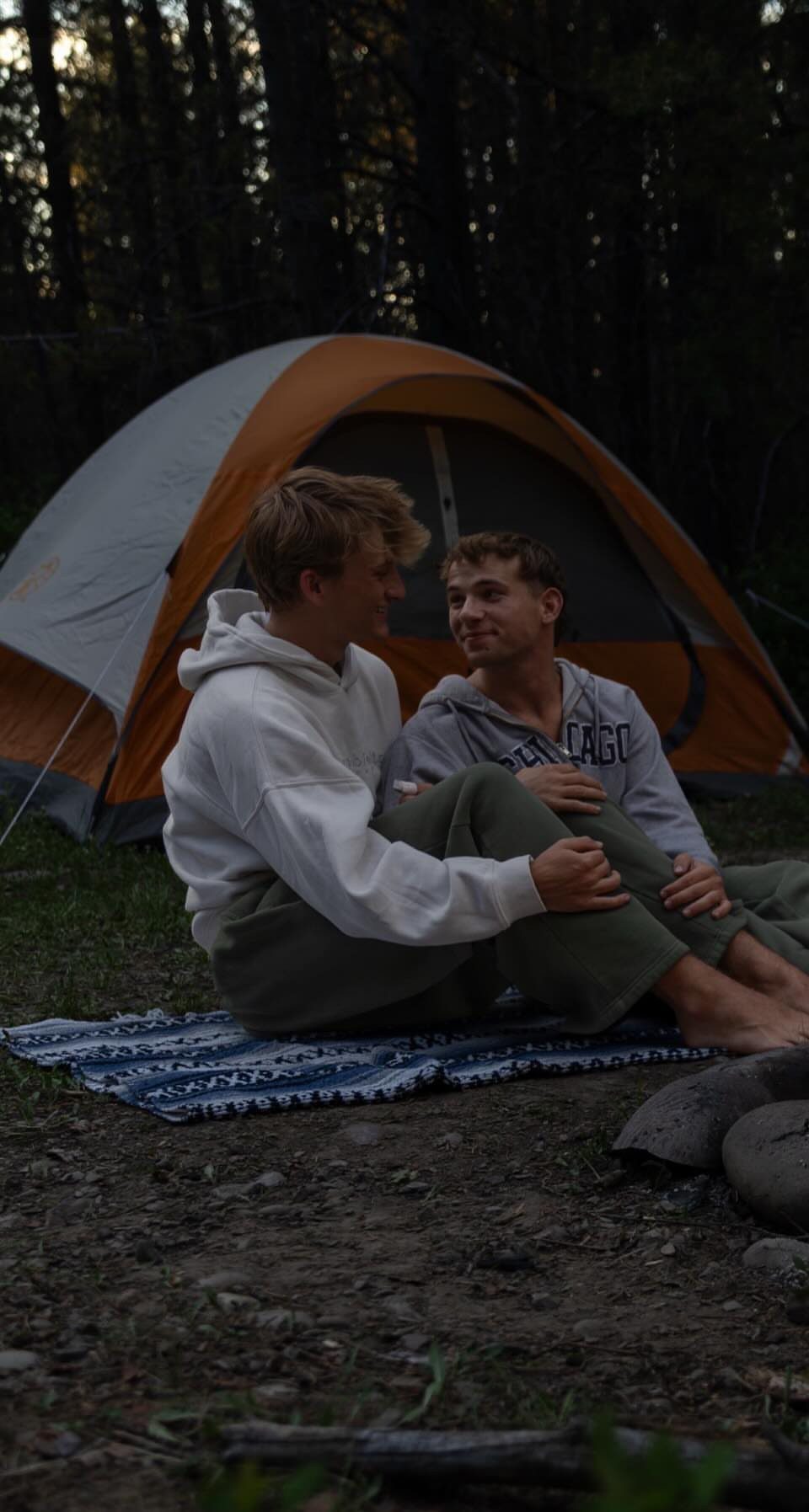 Two teenage boys chatting outside an orange tent in a wooded forest area.