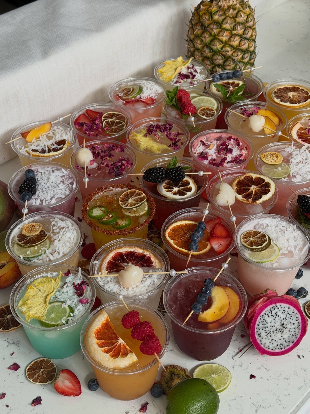 Colorful garnished drinks in plastic cups arranged on a white table with pineapple.
