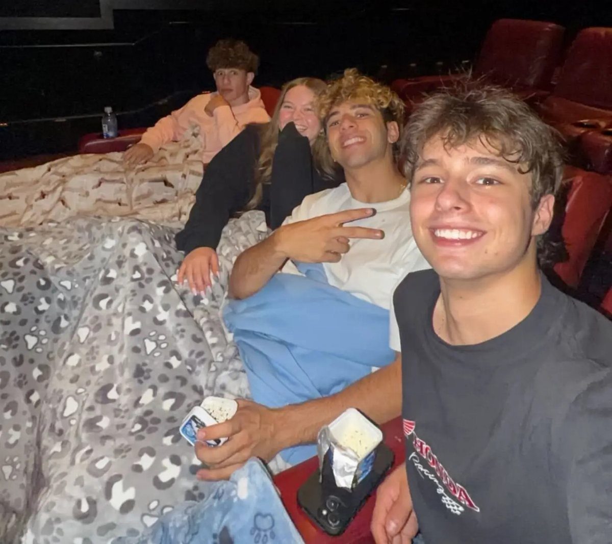 Four teenagers smiling while sitting in a movie theater with blankets and snacks.