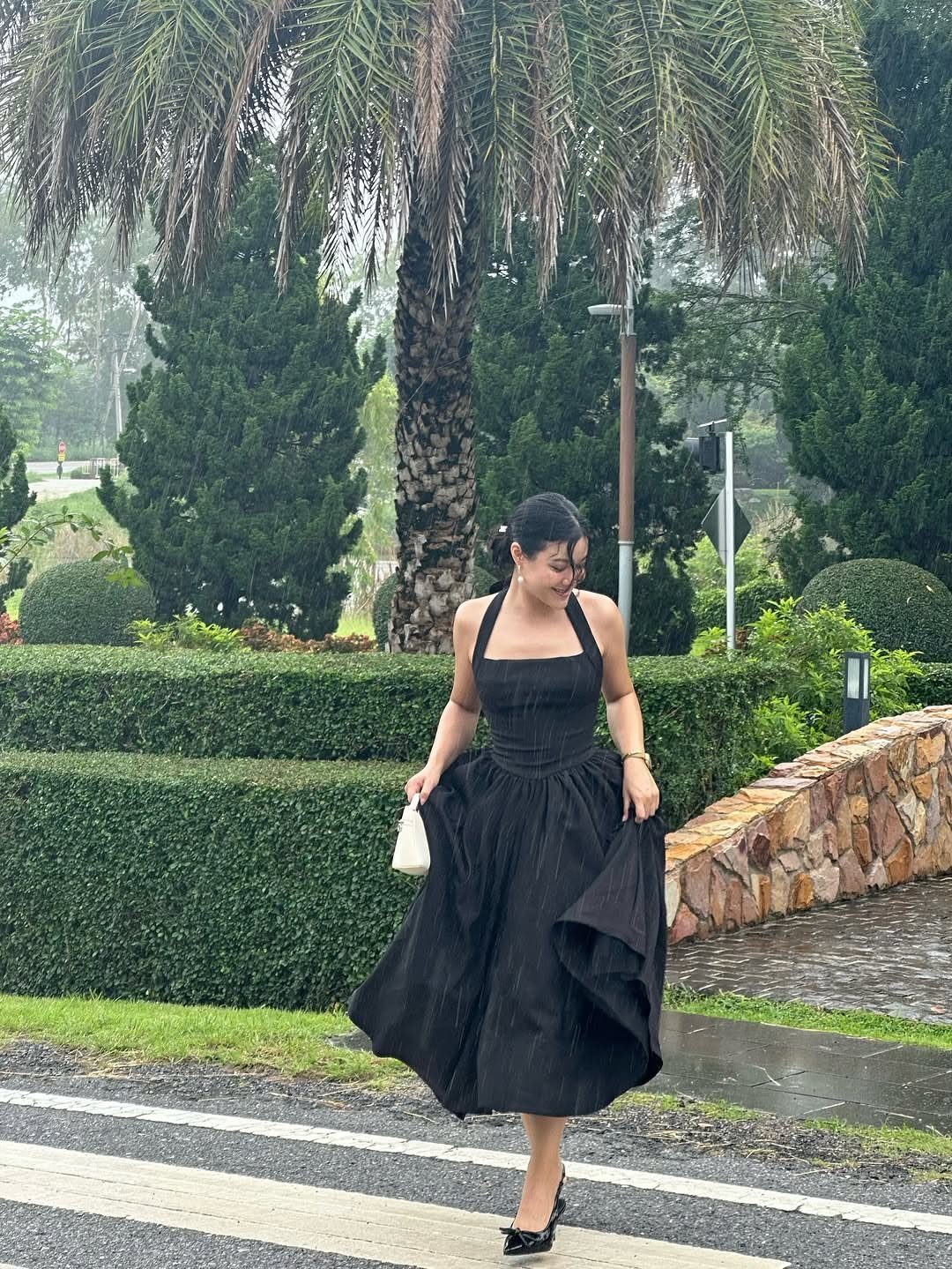 Woman in a black fit-and-flare midi dress walking outdoors near green hedges.