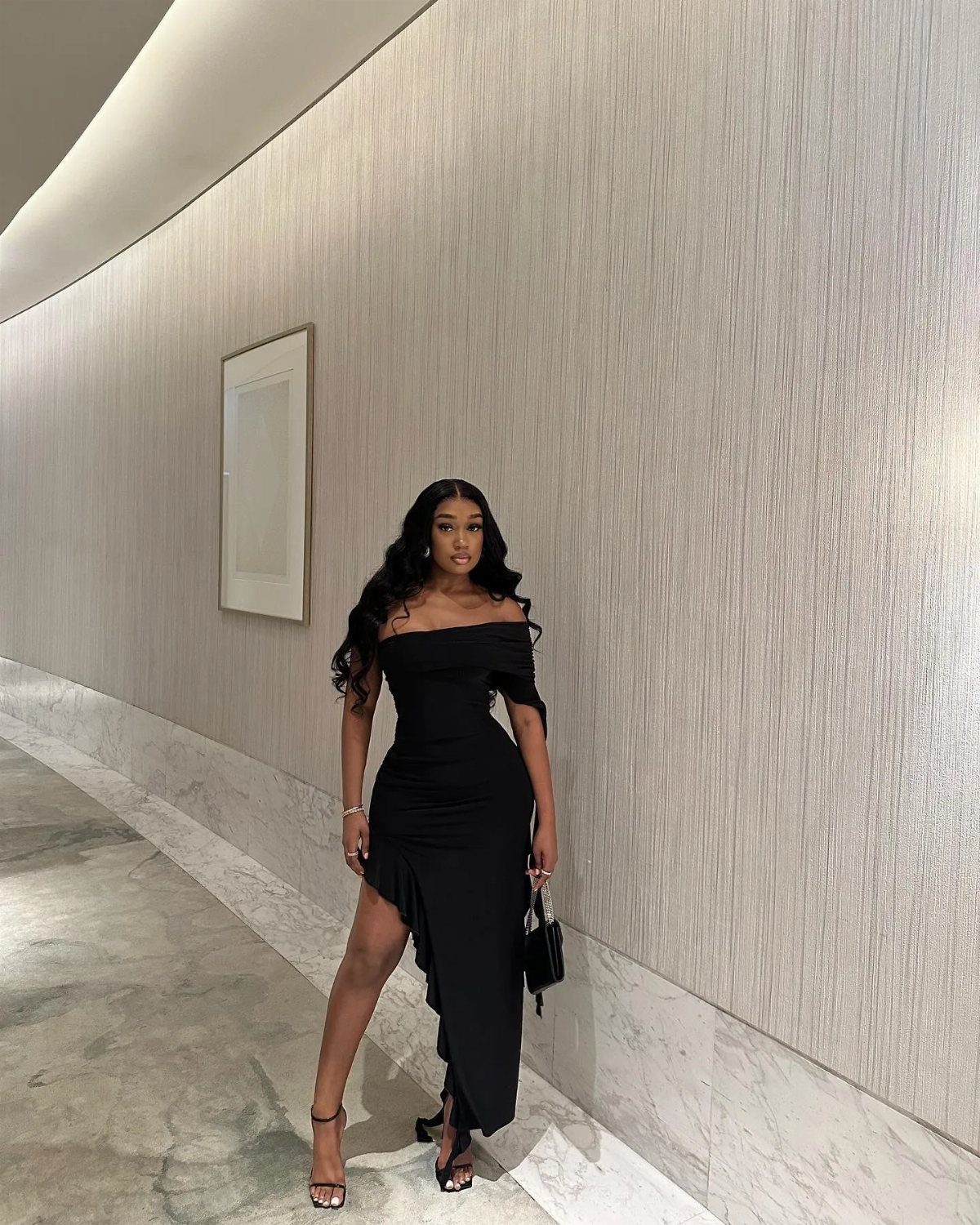 Woman in a long black off-the-shoulder dress with a high slit posing indoors.