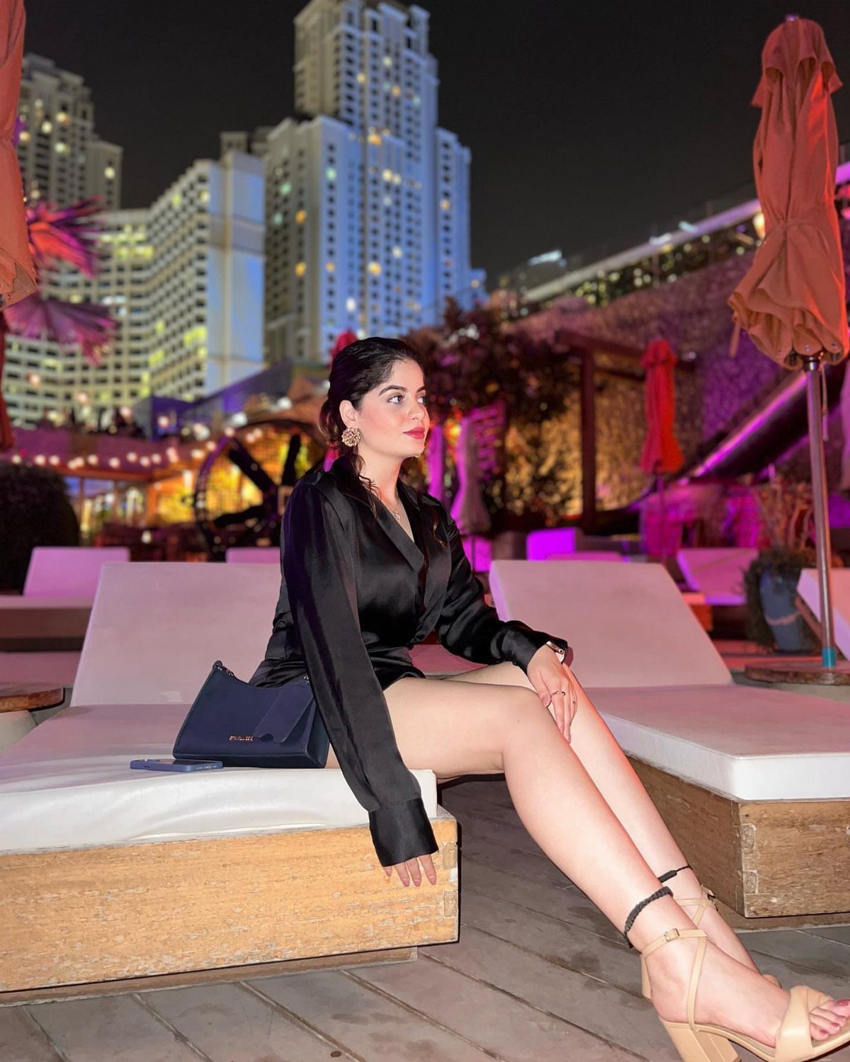 Woman in a black satin mini dress sitting on a lounge chair at night.