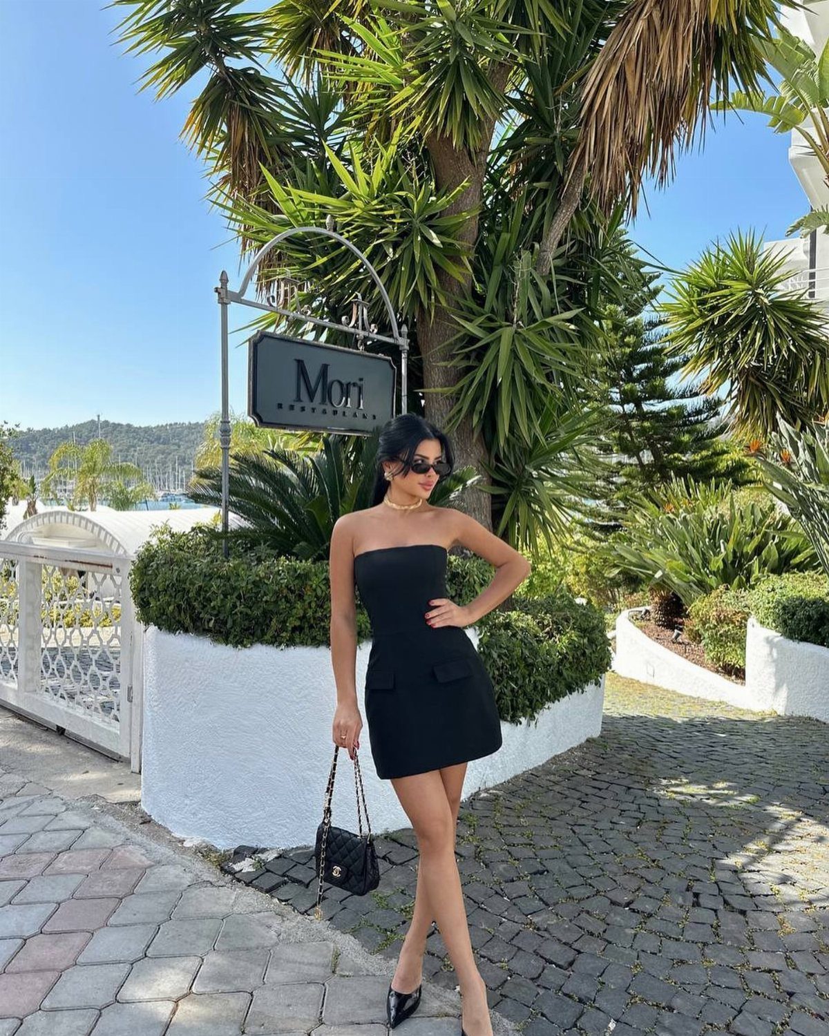 Woman in a black strapless mini dress posing outdoors by palm trees.