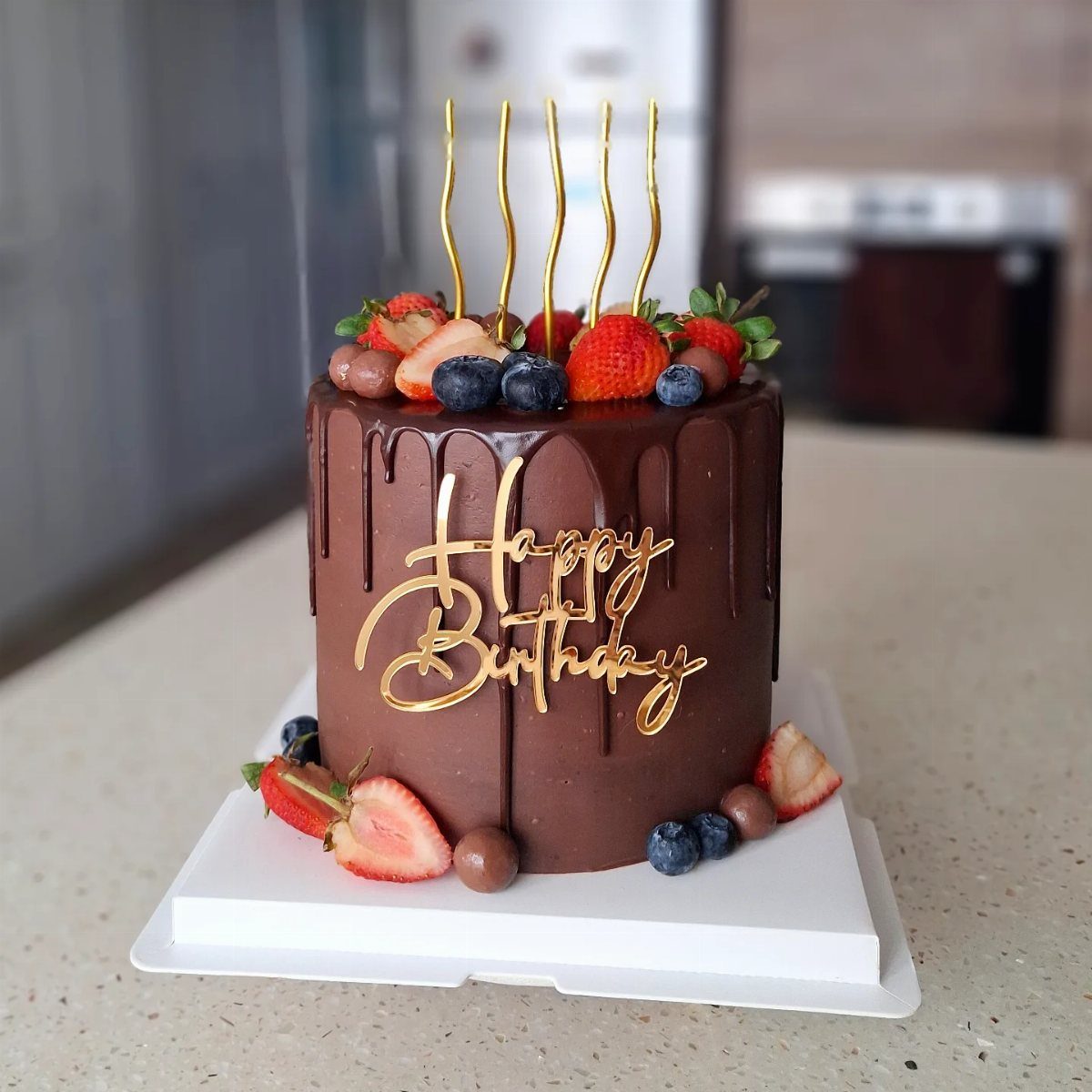 Chocolate drip cake topped with fresh berries, gold candles, and a gold birthday topper.