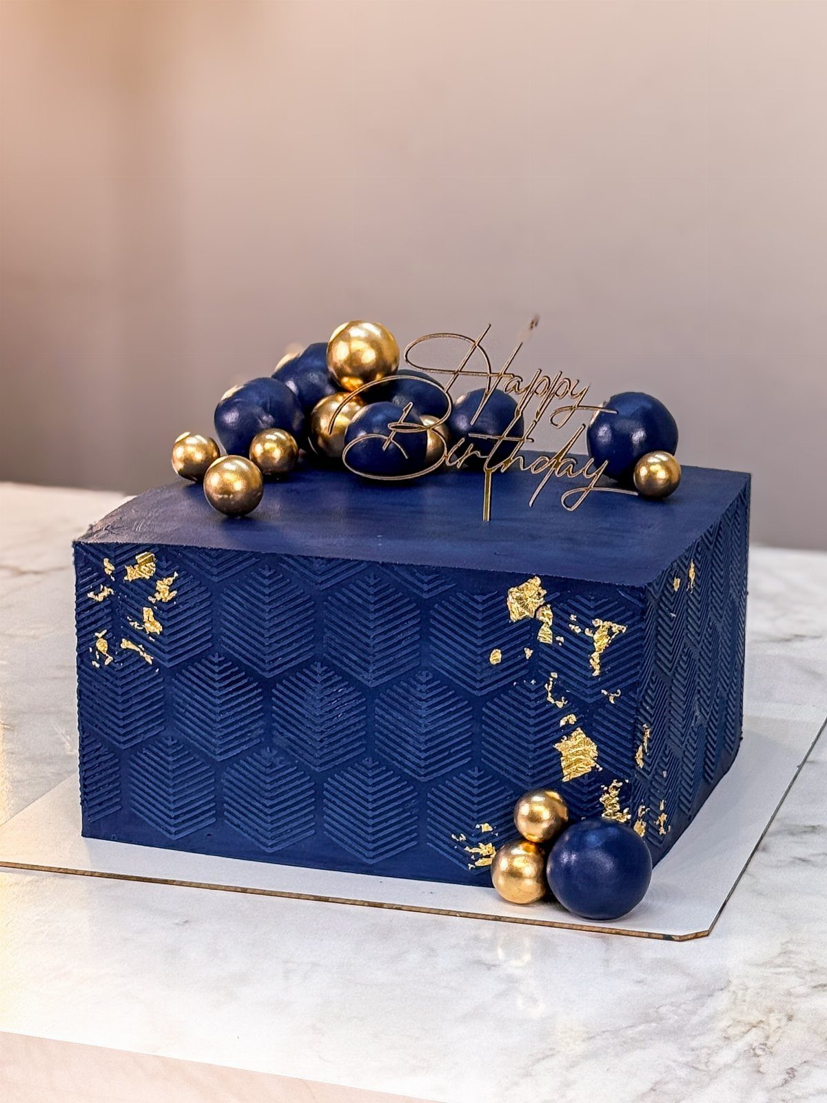 Navy blue rectangular cake with gold leaf, blue and gold spheres, and birthday topper.