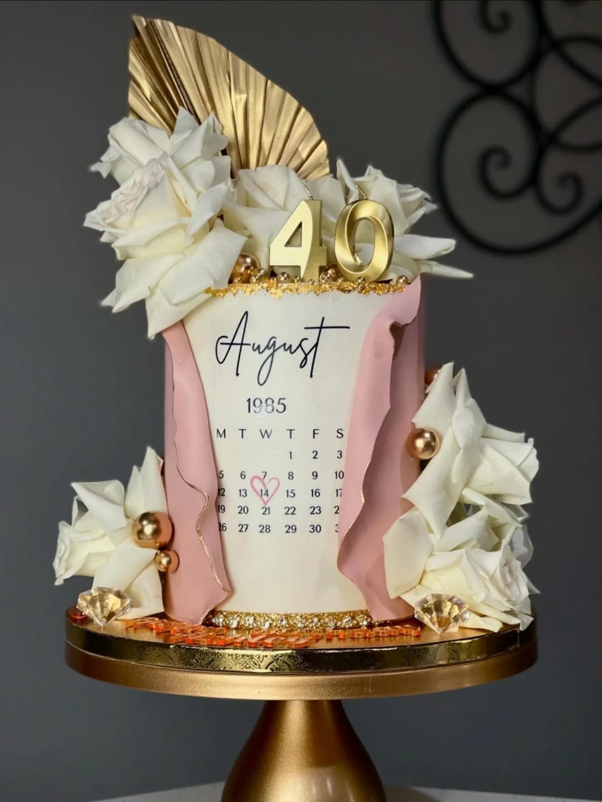 White 40th birthday cake with pink drapes, white roses, and August 1985 calendar.