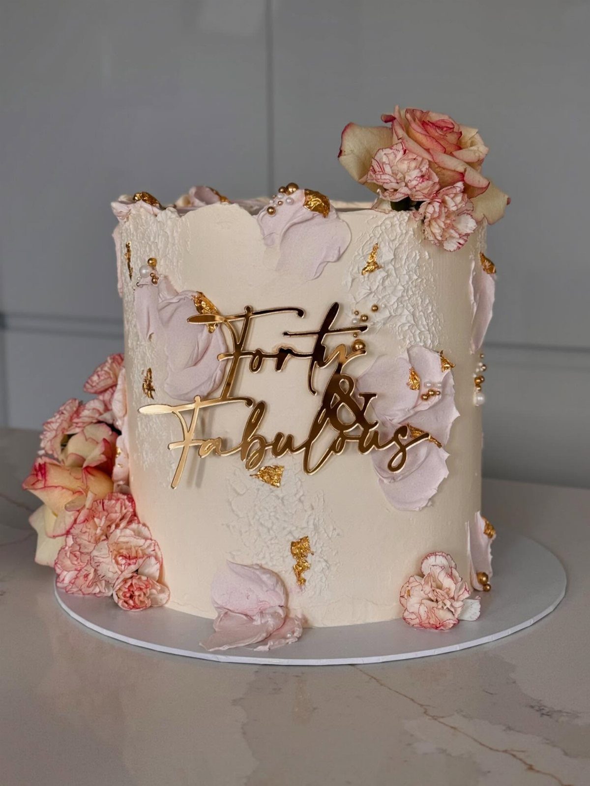 White birthday cake with pink frosting petals, fresh roses, and gold Forty and Fabulous topper