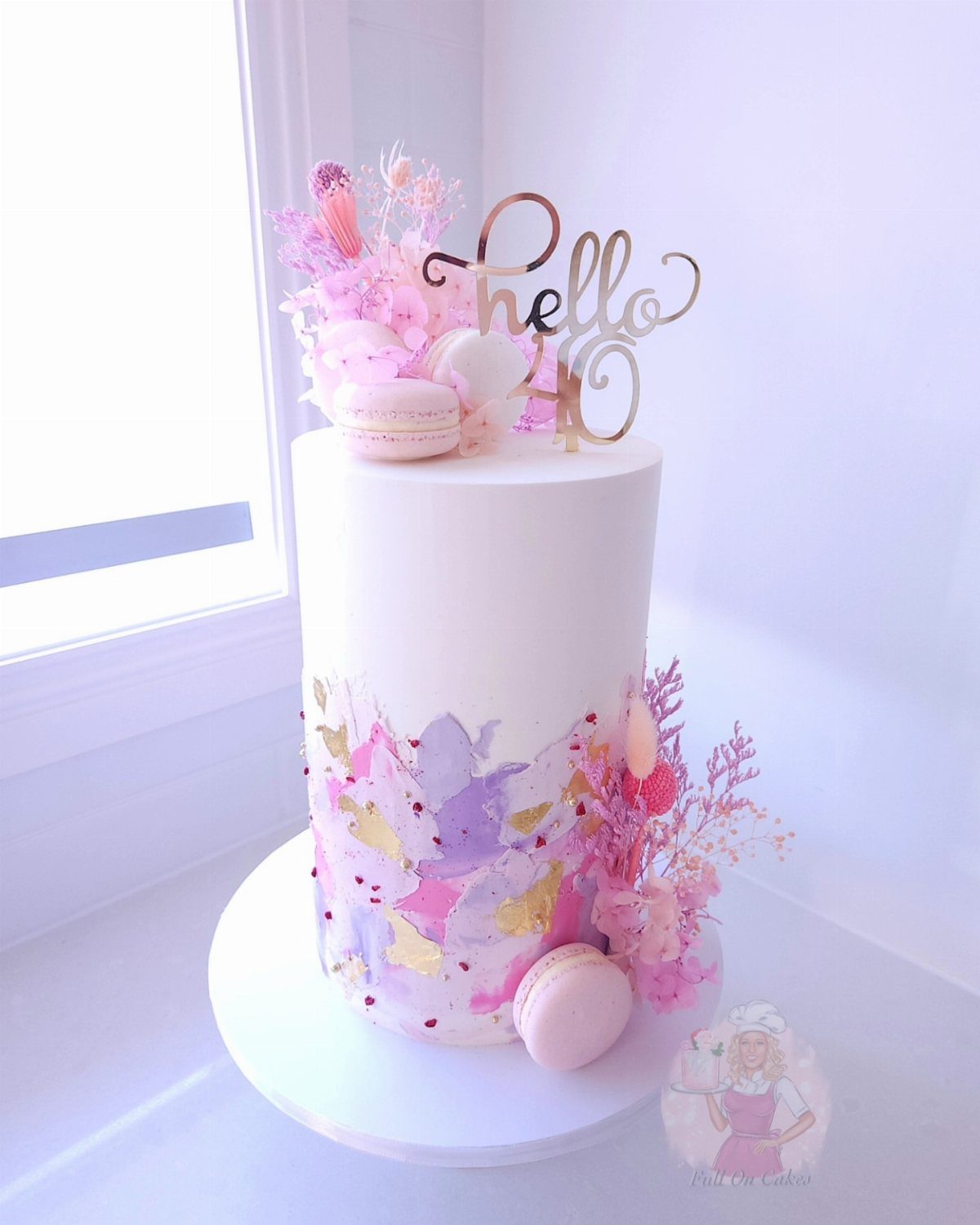 White 40th birthday cake decorated with pink flowers, macarons, and gold topper.