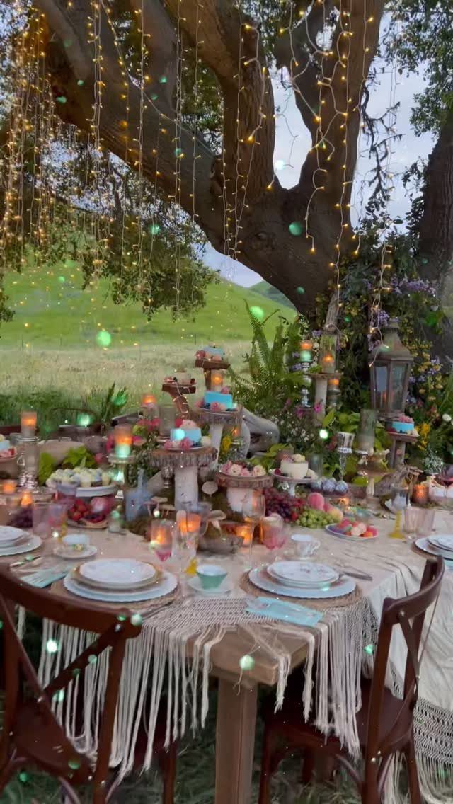 Outdoor dinner table with fairy lights, desserts, and floral greenery under a tree.