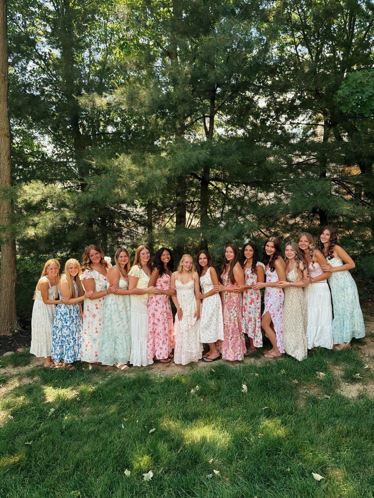 Teenage girls in floral summer dresses posing together under large green trees outdoors.