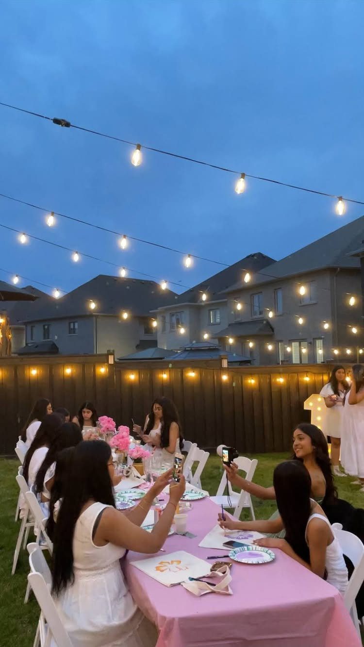 Young women in white dresses dining at a pink table under outdoor string lights.