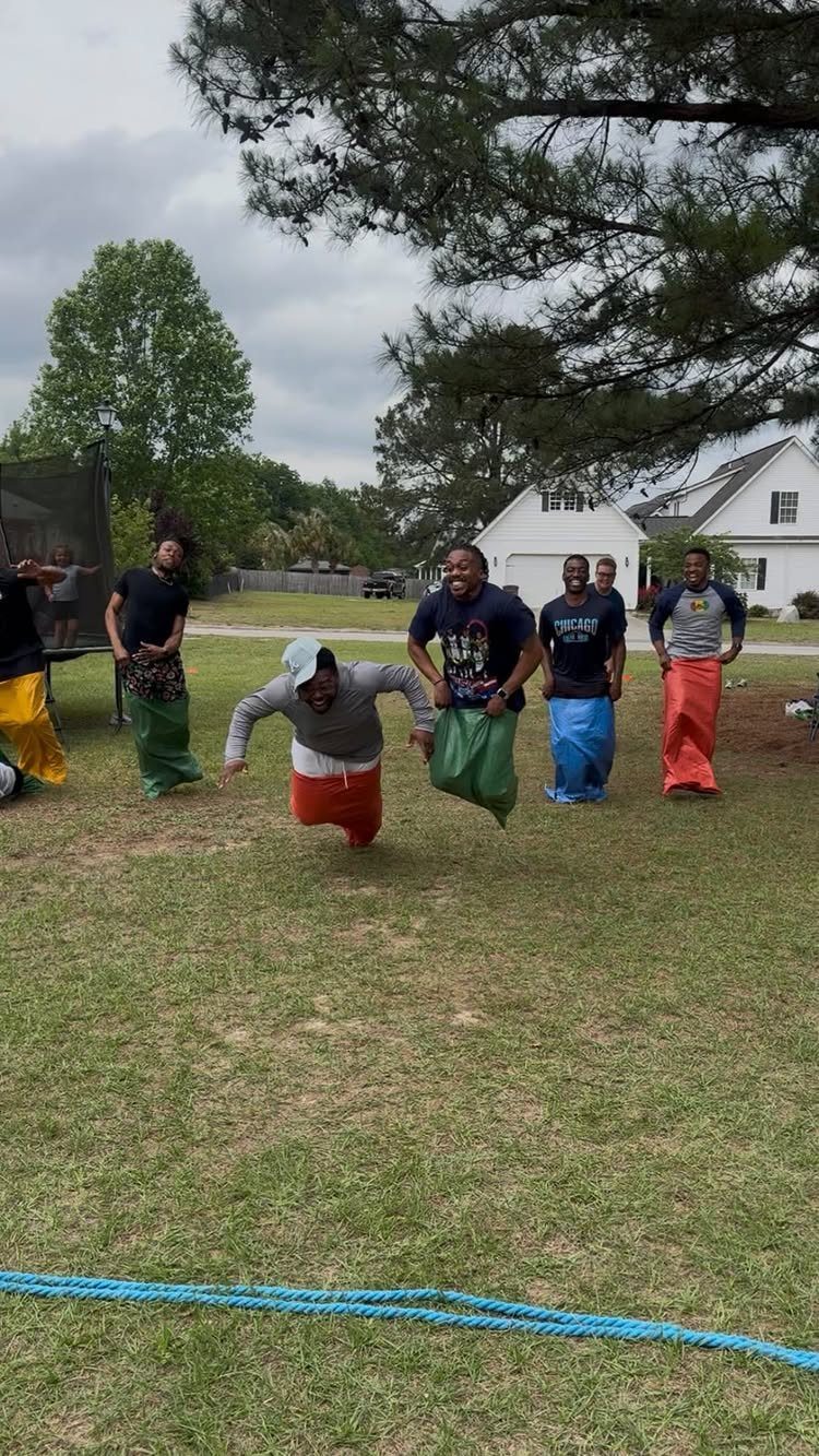 Men jumping in colorful sacks during a potato sack race on a lawn.