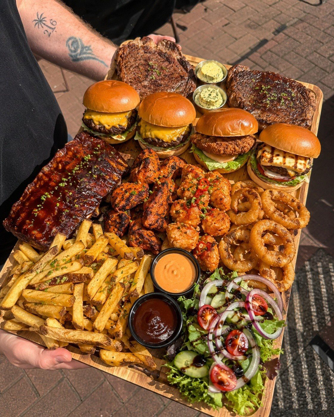 Wooden platter filled with sliders, ribs, steaks, fries, and salad held by a person.