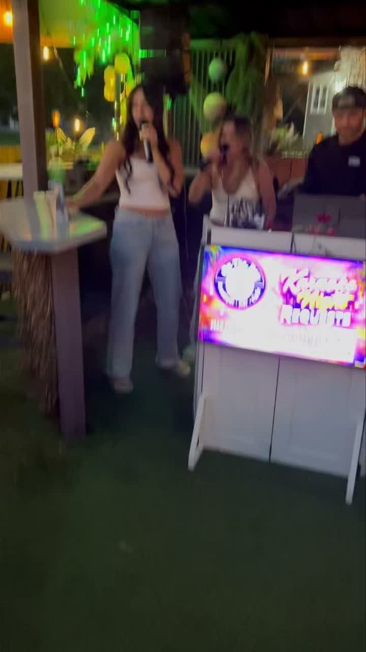 Two young women singing karaoke at an outdoor night party with a DJ booth.