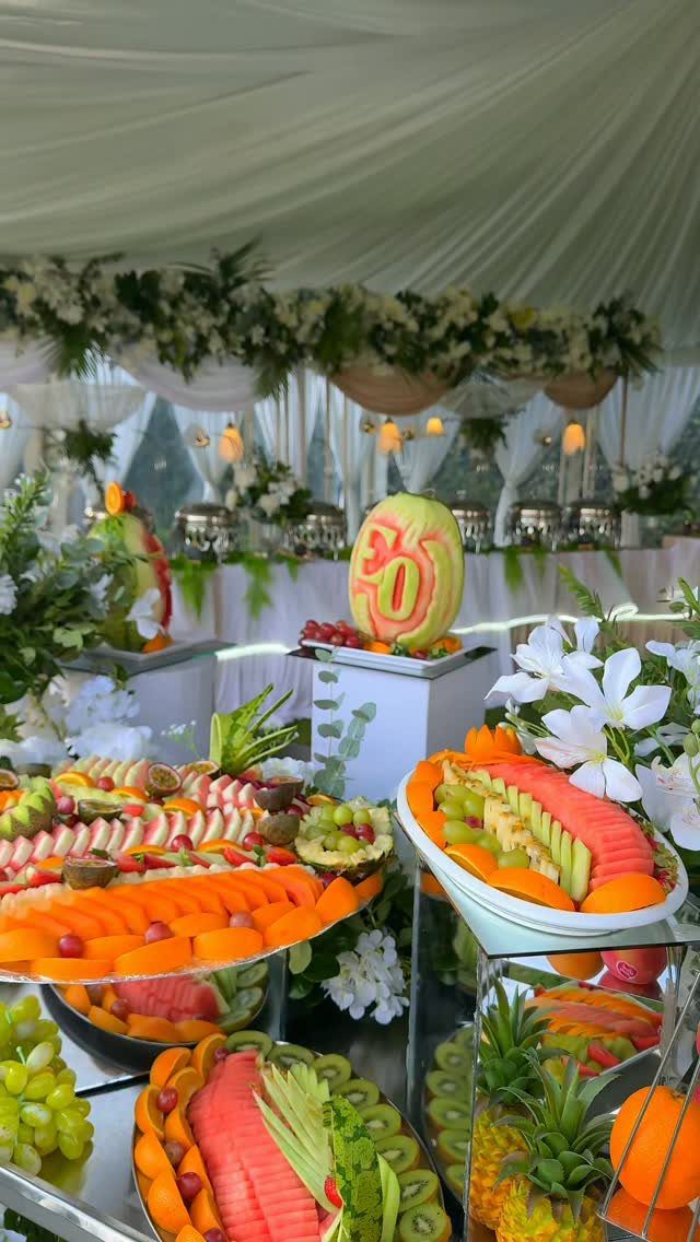 Fruit buffet with carved melon and sliced tropical fruits under a white tent.