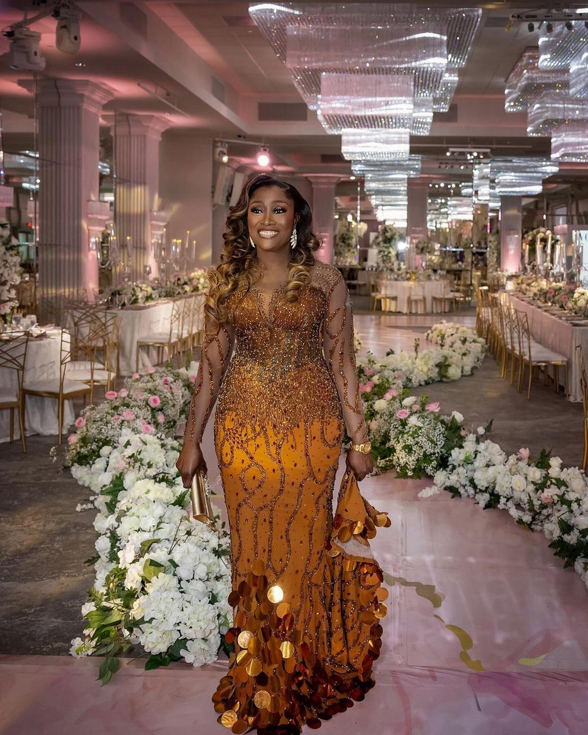 Woman in a beaded orange gown standing in a decorated luxury ballroom.