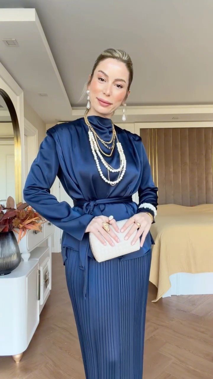 Woman in navy blue satin blouse and pleated skirt with pearl jewelry and clutch.