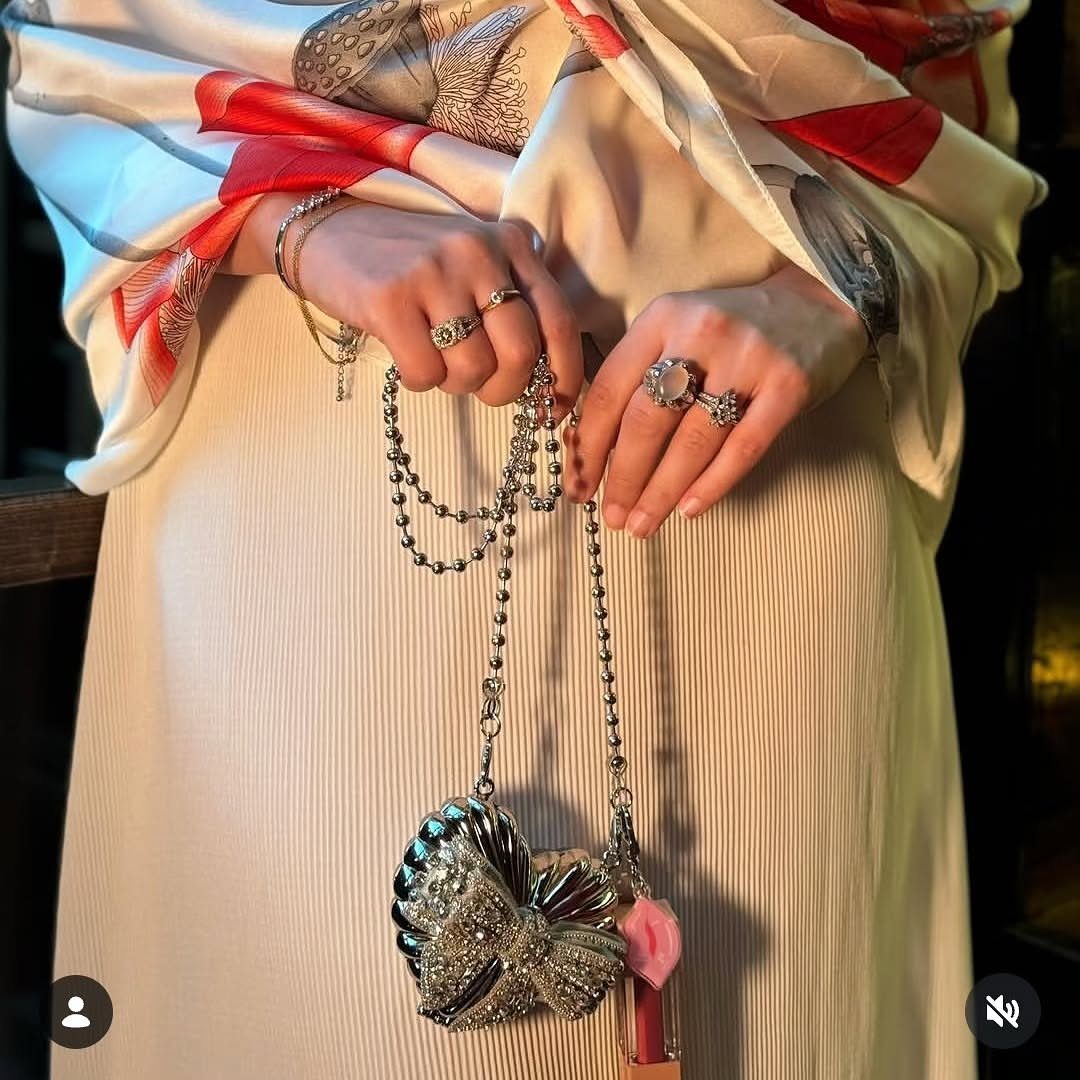 Woman in cream pleated dress and red patterned scarf holding a silver bow handbag