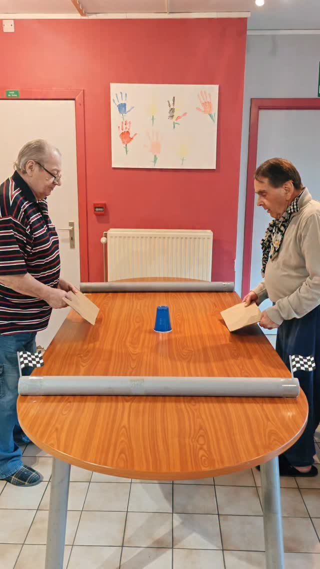Two seniors playing a sliding cup game on a wooden table in a room.