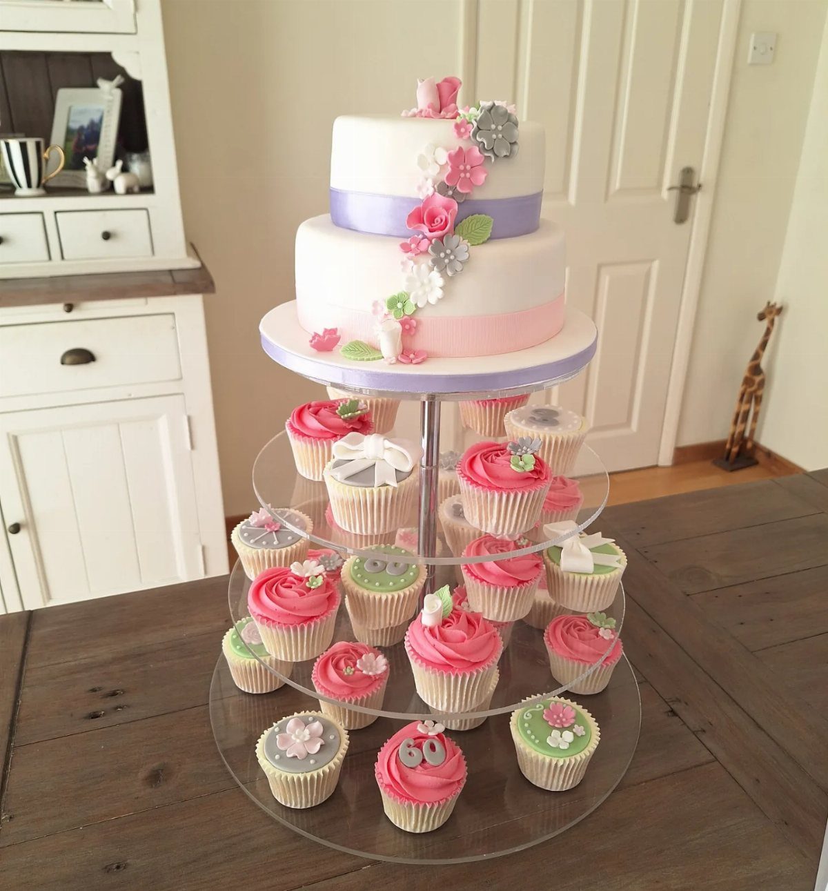 Two-tier white floral cake atop a clear stand filled with colorful cupcakes.