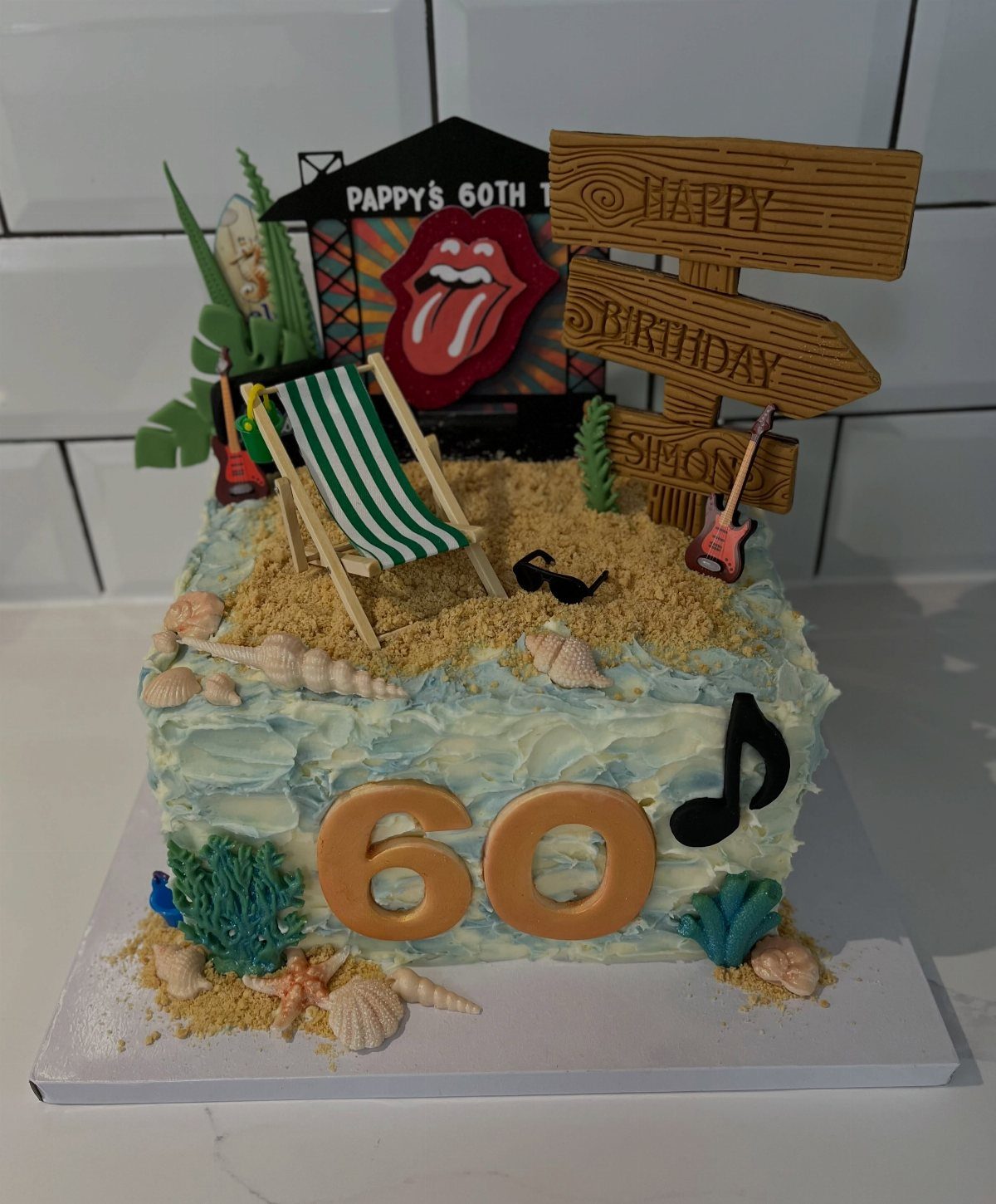 Beach-themed 60th birthday cake with blue frosting, sand, seashells, and miniature accessories.