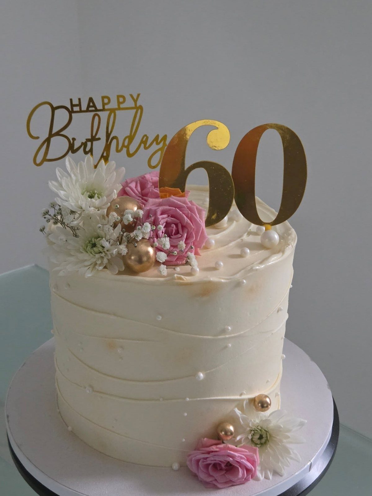 White frosted birthday cake decorated with pink roses, white daisies, and gold 60 topper.