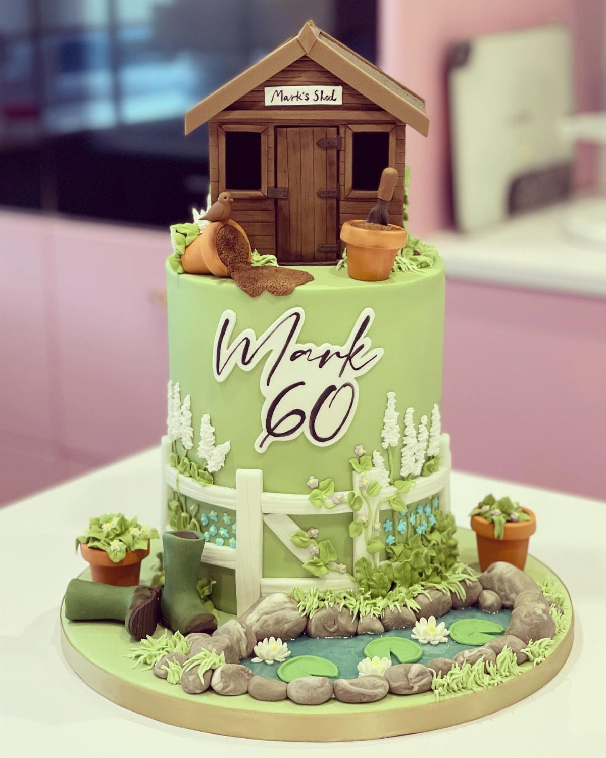 Green 60th birthday cake decorated with a garden shed, pond, and edible tools.