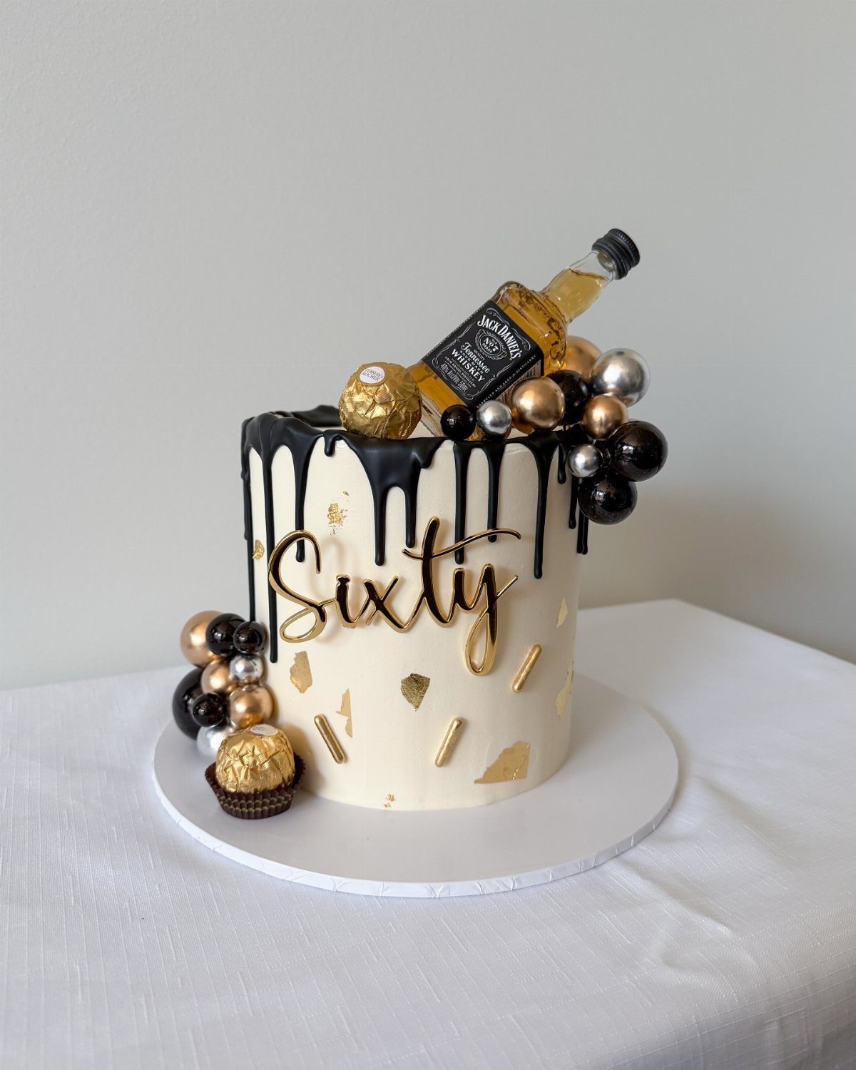 White birthday cake with black chocolate drip, gold Sixty lettering, and mini liquor bottle.