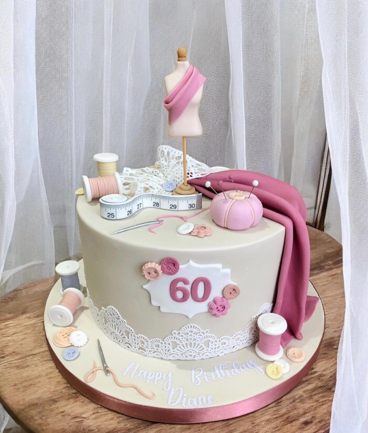 Beige 60th birthday cake decorated with sewing tools, a mannequin, and pink fabric.