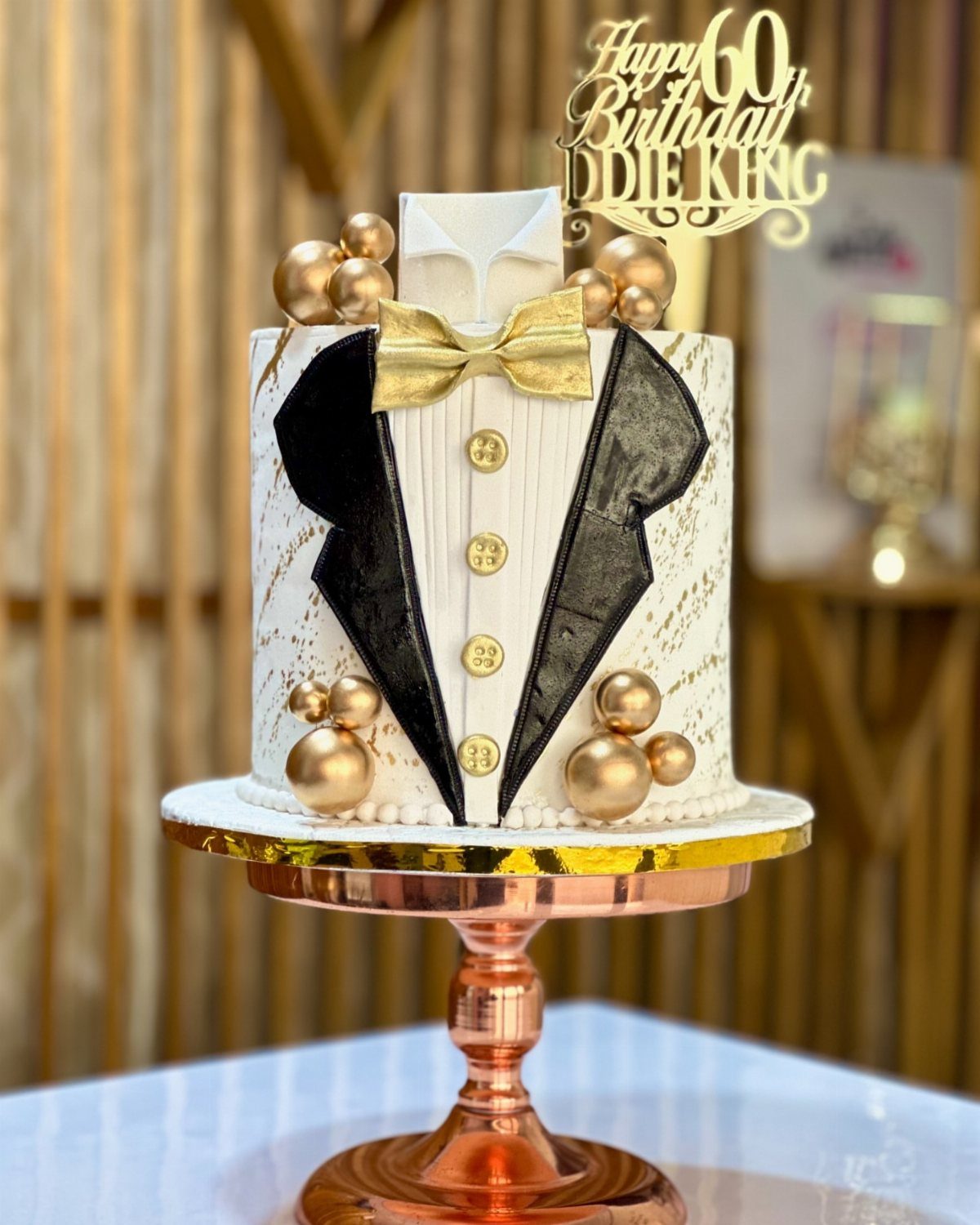 Tuxedo themed 60th birthday cake with gold accents on a copper stand