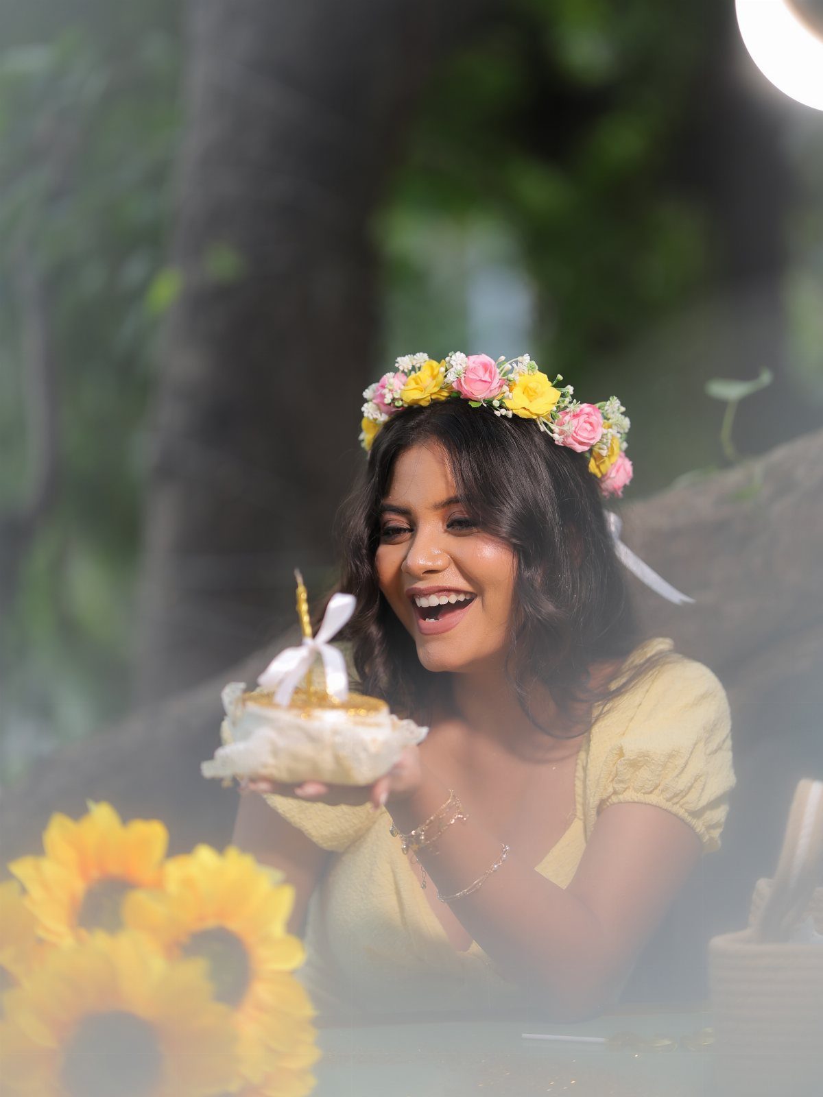 Woman in yellow dress and floral crown holding a small birthday cake outdoors.