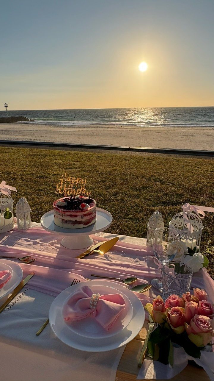 Beachside picnic table with a cake, pink roses, and lanterns at sunset.