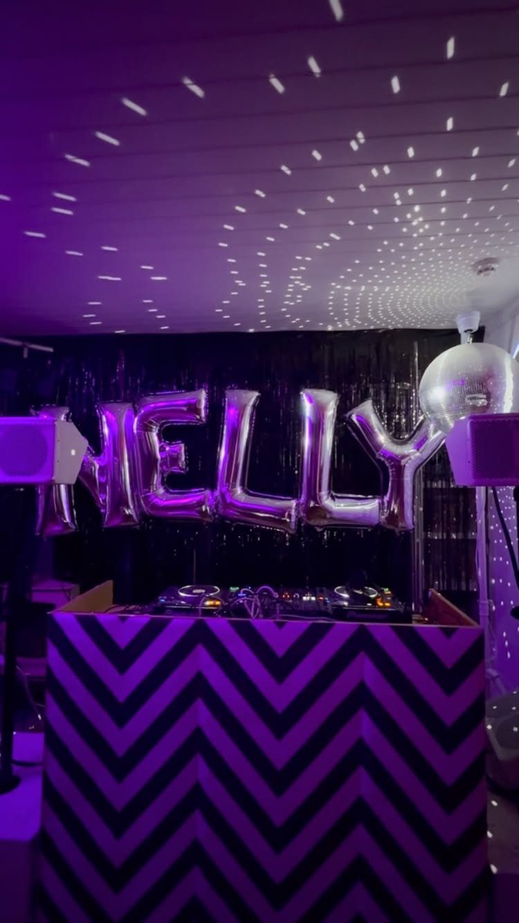 Party setup with purple lighting, silver balloons spelling NELLY, and a chevron DJ booth.