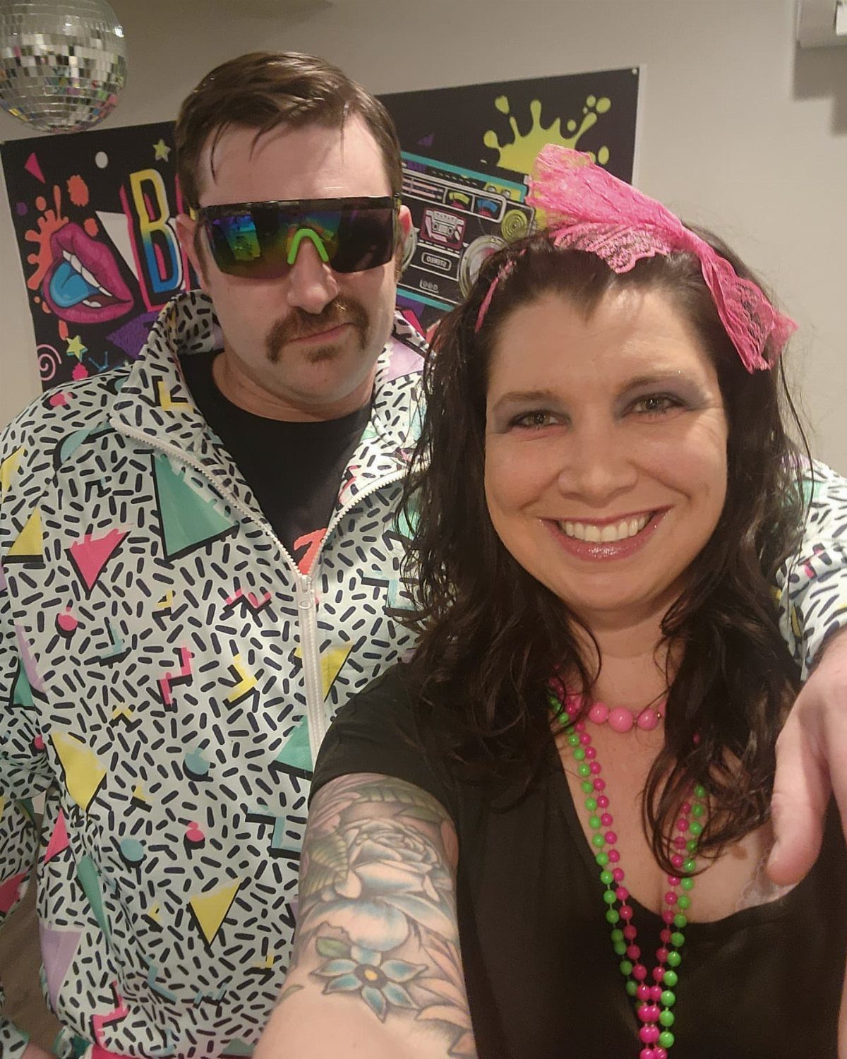 Man in 80s geometric jacket and woman with pink headband posing together.