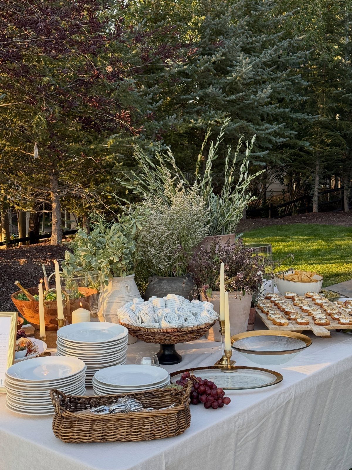 Outdoor buffet table with white plates, appetizers, and floral arrangements in a garden.