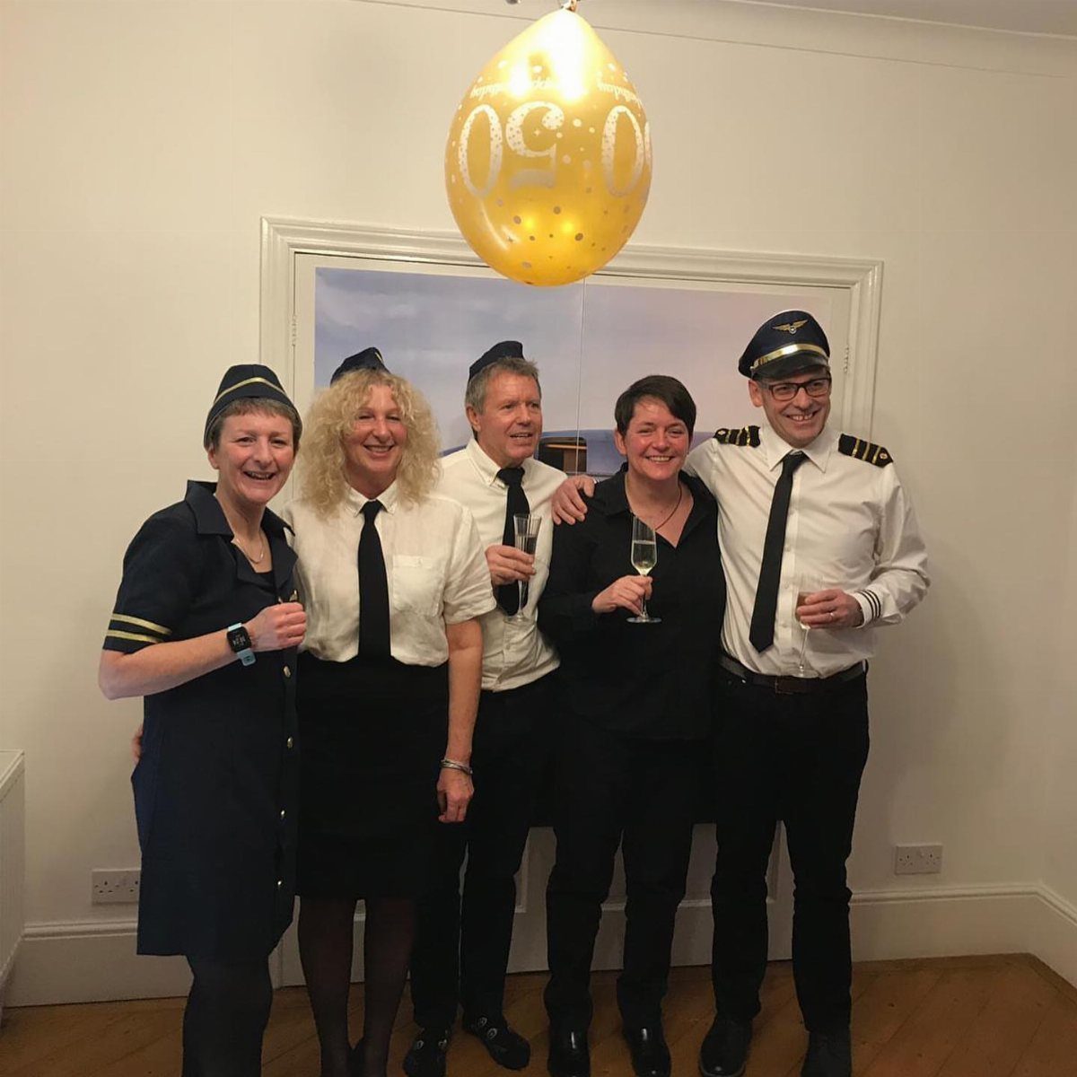 Five people in pilot and flight attendant costumes posing with champagne and a 50 balloon.