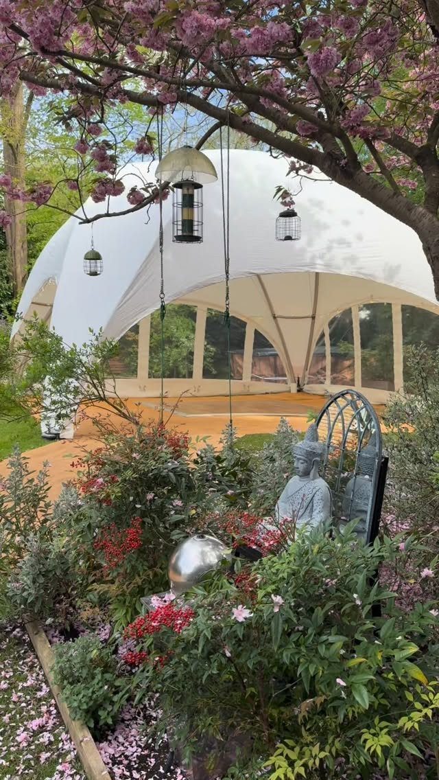 White dome event tent in a lush garden with pink blossoms and lanterns.
