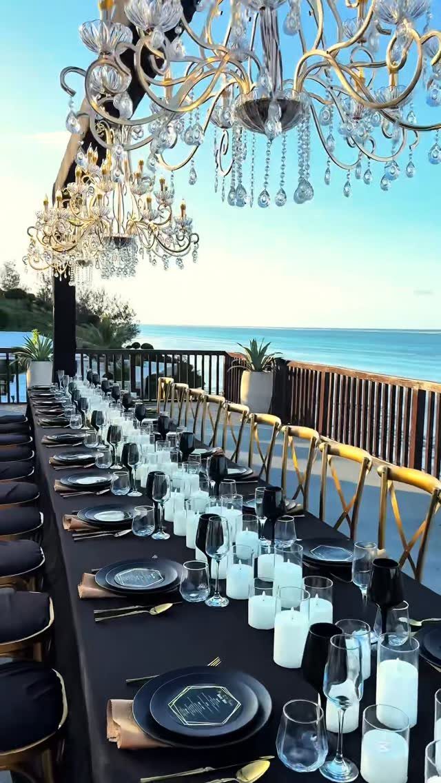 Elegant outdoor dinner table with black linens, gold accents, and crystal chandeliers by the ocean.