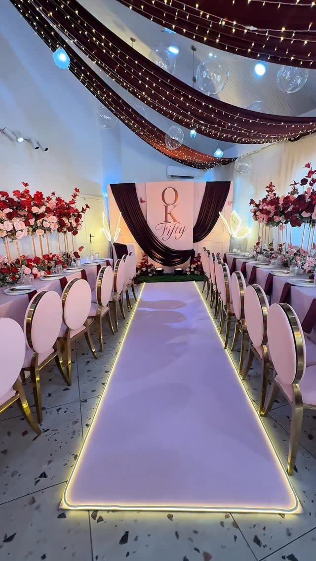 Elegant party venue with pink aisle, gold chairs, and red floral centerpieces.